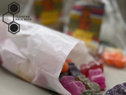 A white paper bag filled with colorful resin jelly babies on a white surface, with 'Cloister Bell Boys' logo in the upper left corner.